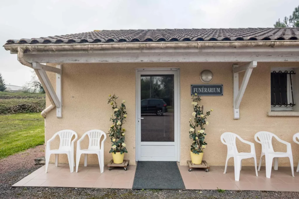 Entrée du funérarium avec porte vitrée, chaises blanches et rosiers en pot.