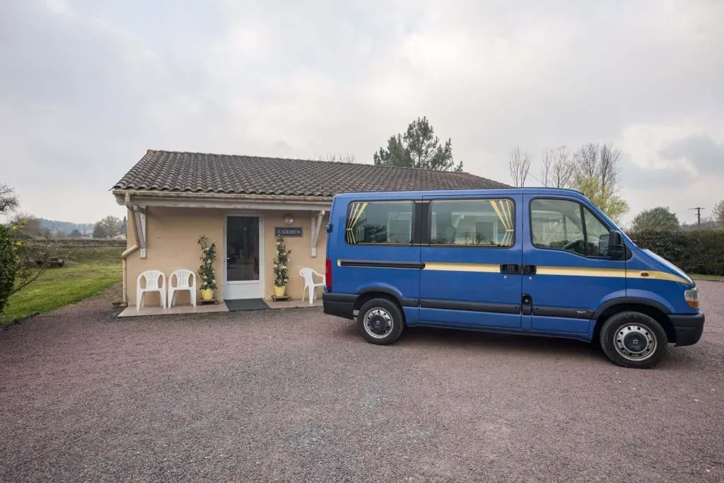 Vue de profil du véhicule funéraire bleu devant l'entrée du funérarium.